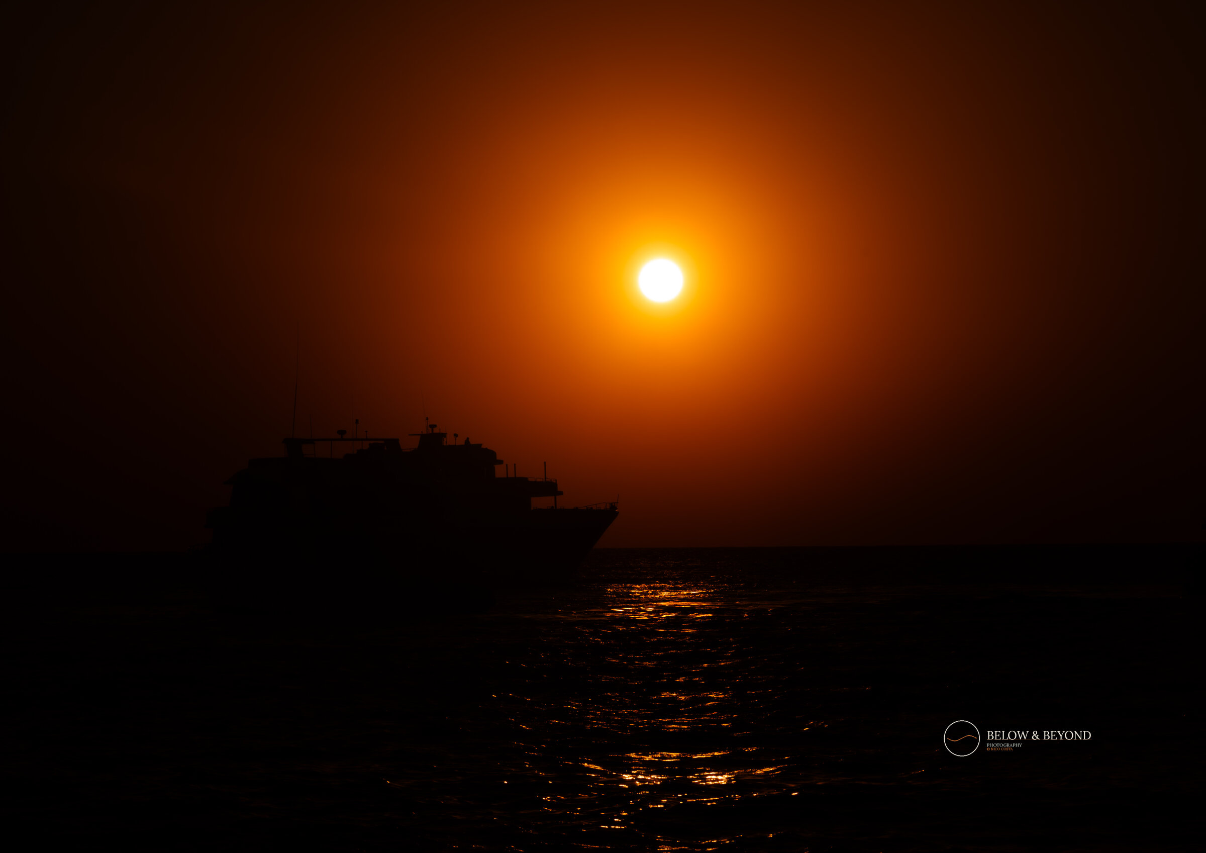 Silhouette of a dive boat against an intense orange-red sunset over calm open water